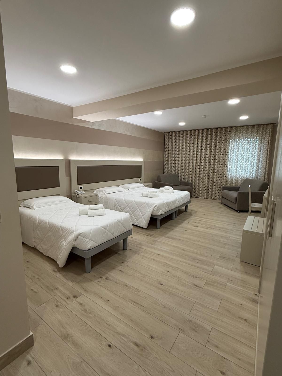 Camera d'albergo con due letti, zona soggiorno e pavimento in legno. Colori neutri.