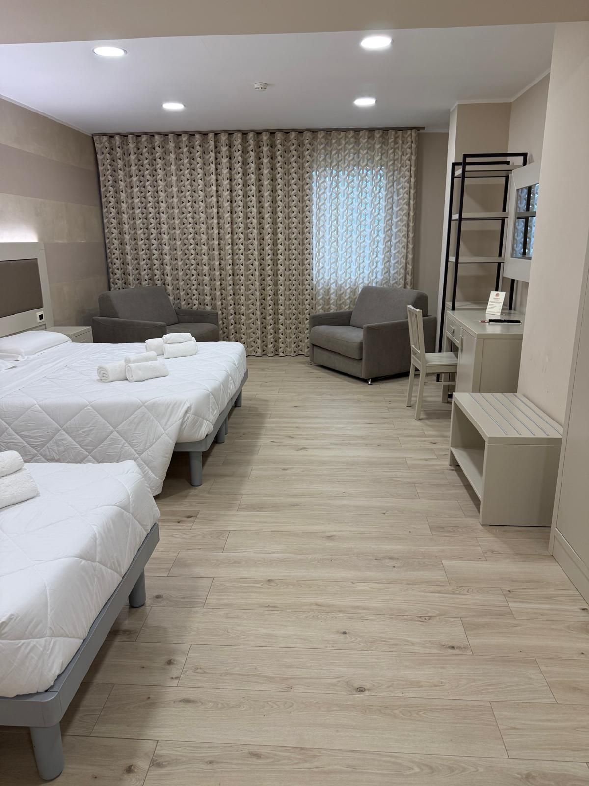 Camera d'albergo con tre letti, due poltrone, scrivania e una finestra con tende a fantasia.