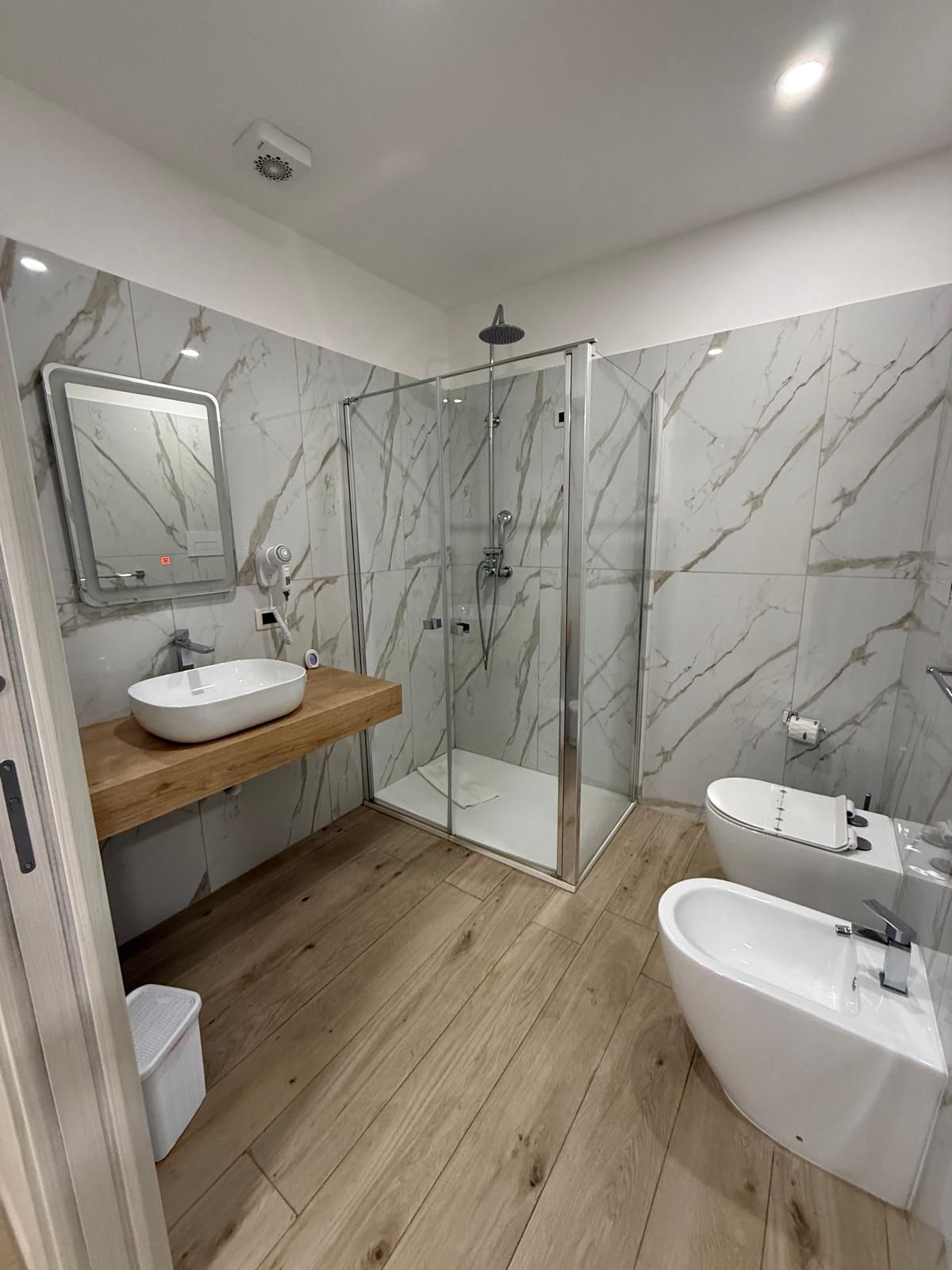 Bagno moderno con piastrelle effetto marmo, doccia in vetro, mobile lavabo in legno e bidet.