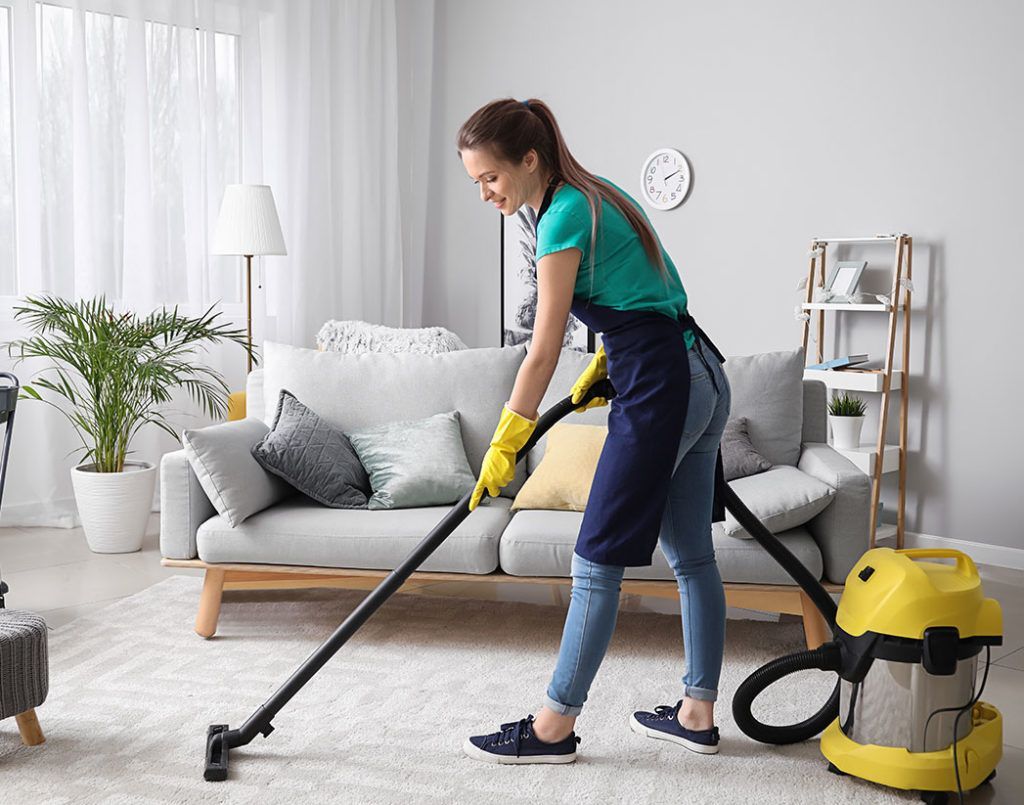 A person in a turquoise shirt and apron vacuums a light-colored rug in a bright, modern living room.