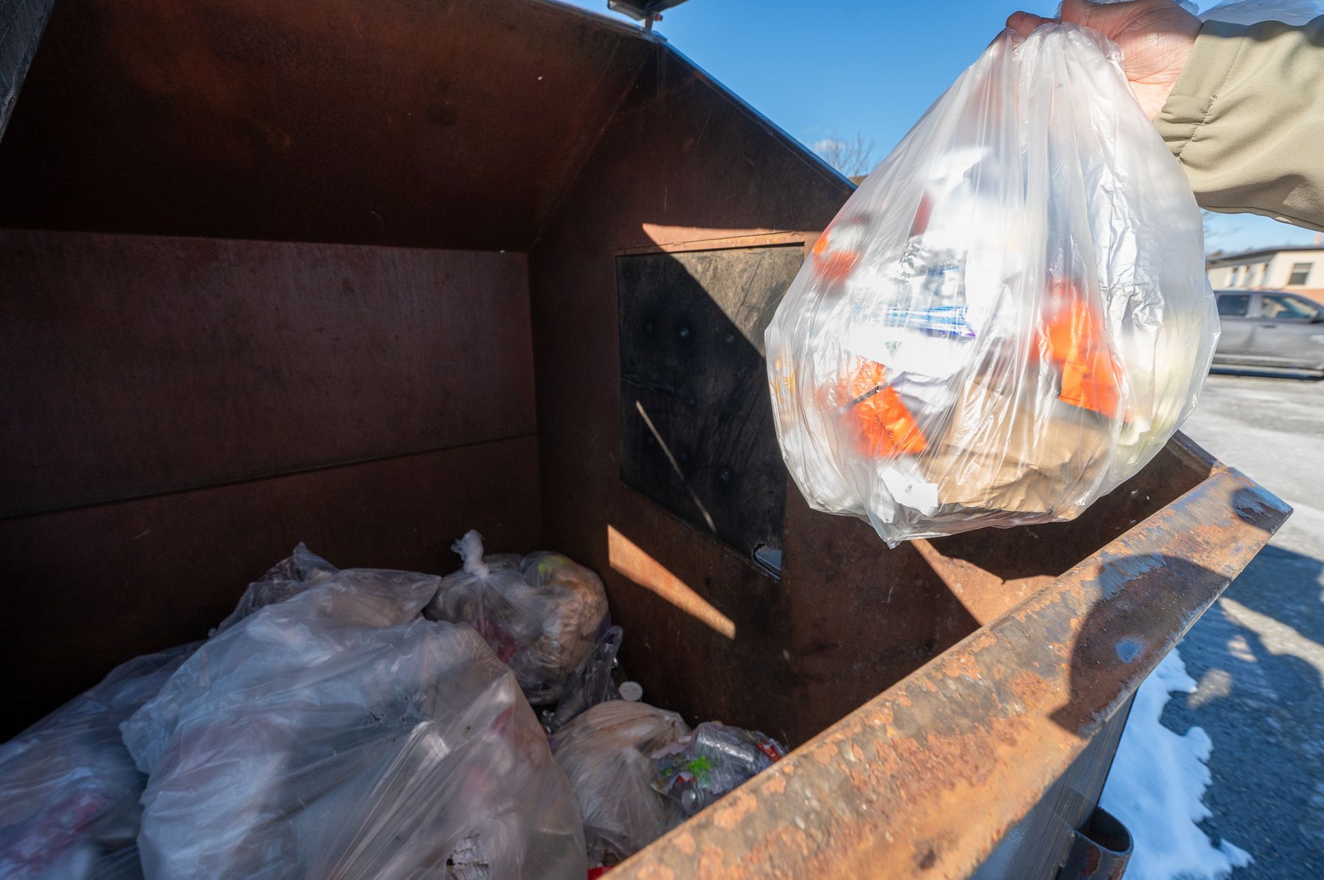 A hand holds a clear plastic bag filled with refuse over a rusted, partially filled outdoor trash dumpster.