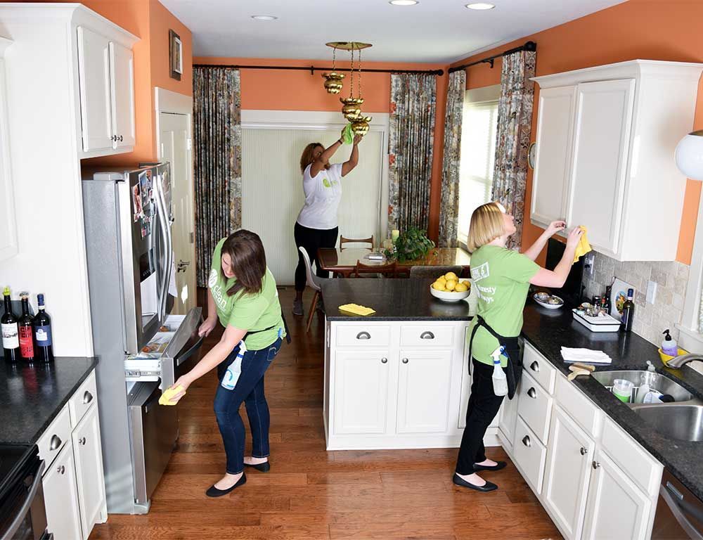 Three cleaners in green uniforms tidy a kitchen, wiping down the refrigerator, light fixtures, and upper cabinets.