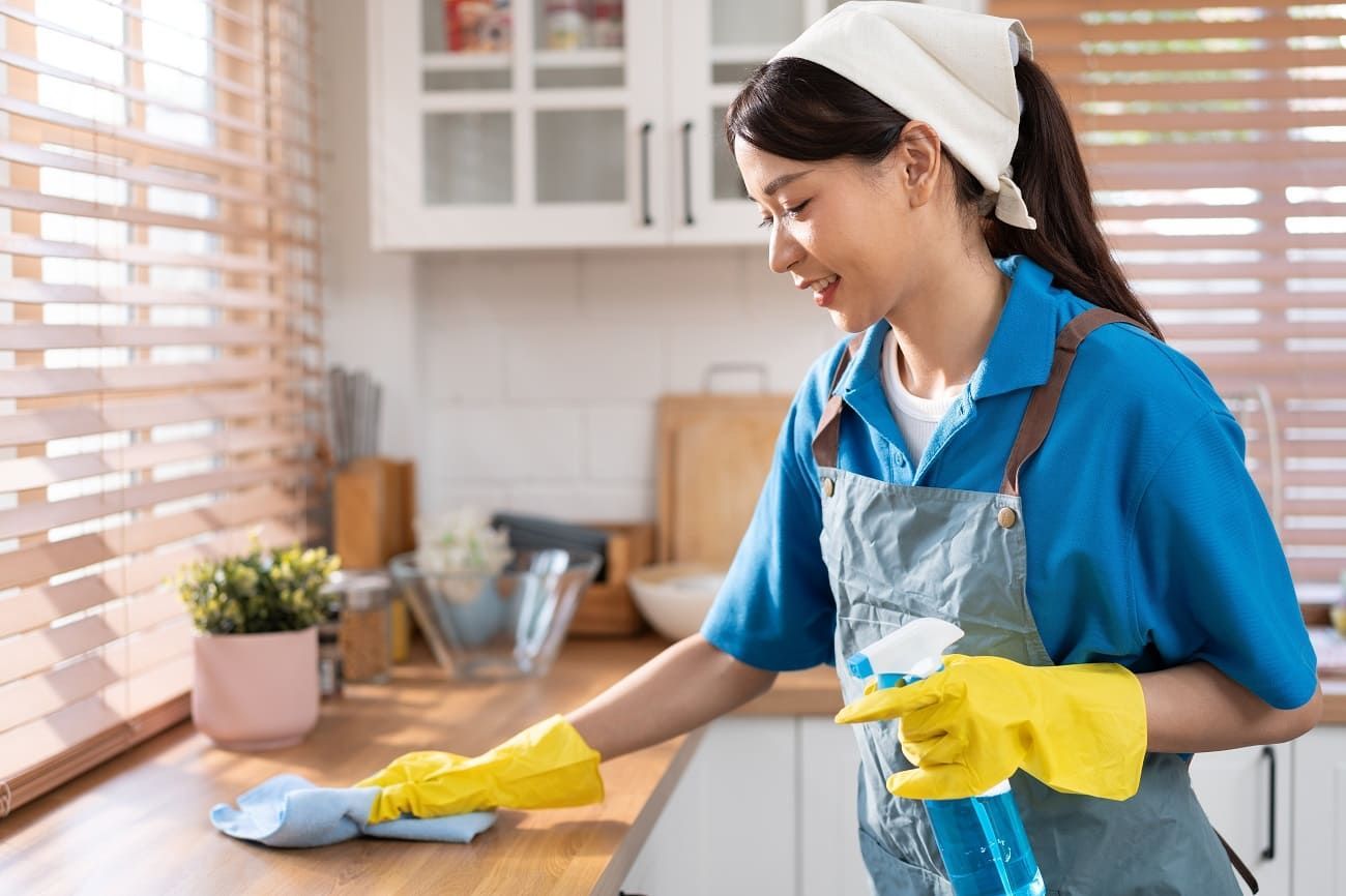 A person in a blue shirt, gray apron, and yellow gloves wipes a wooden kitchen countertop with a cleaning cloth.
