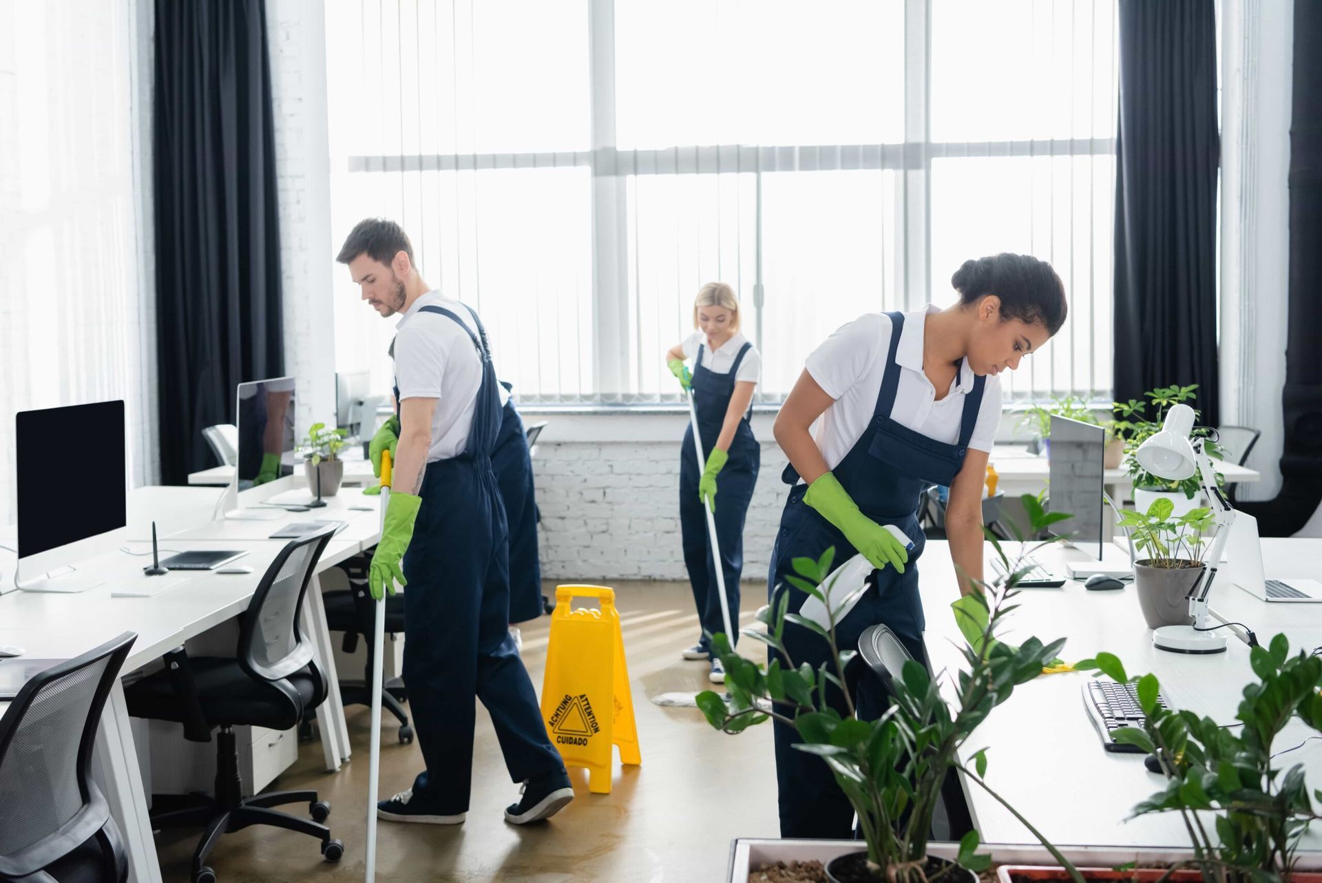 Three professional cleaners in uniforms and green gloves work in a modern office with desks and plants.