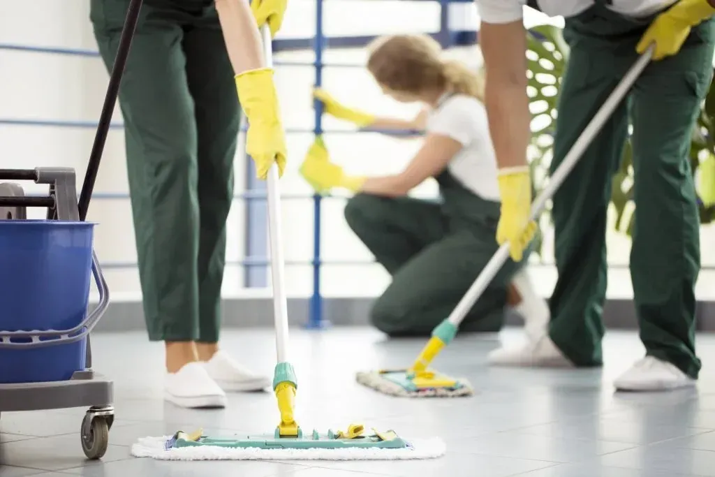 Professional cleaners in green uniforms and yellow gloves mop a floor while another person cleans a railing in the back.