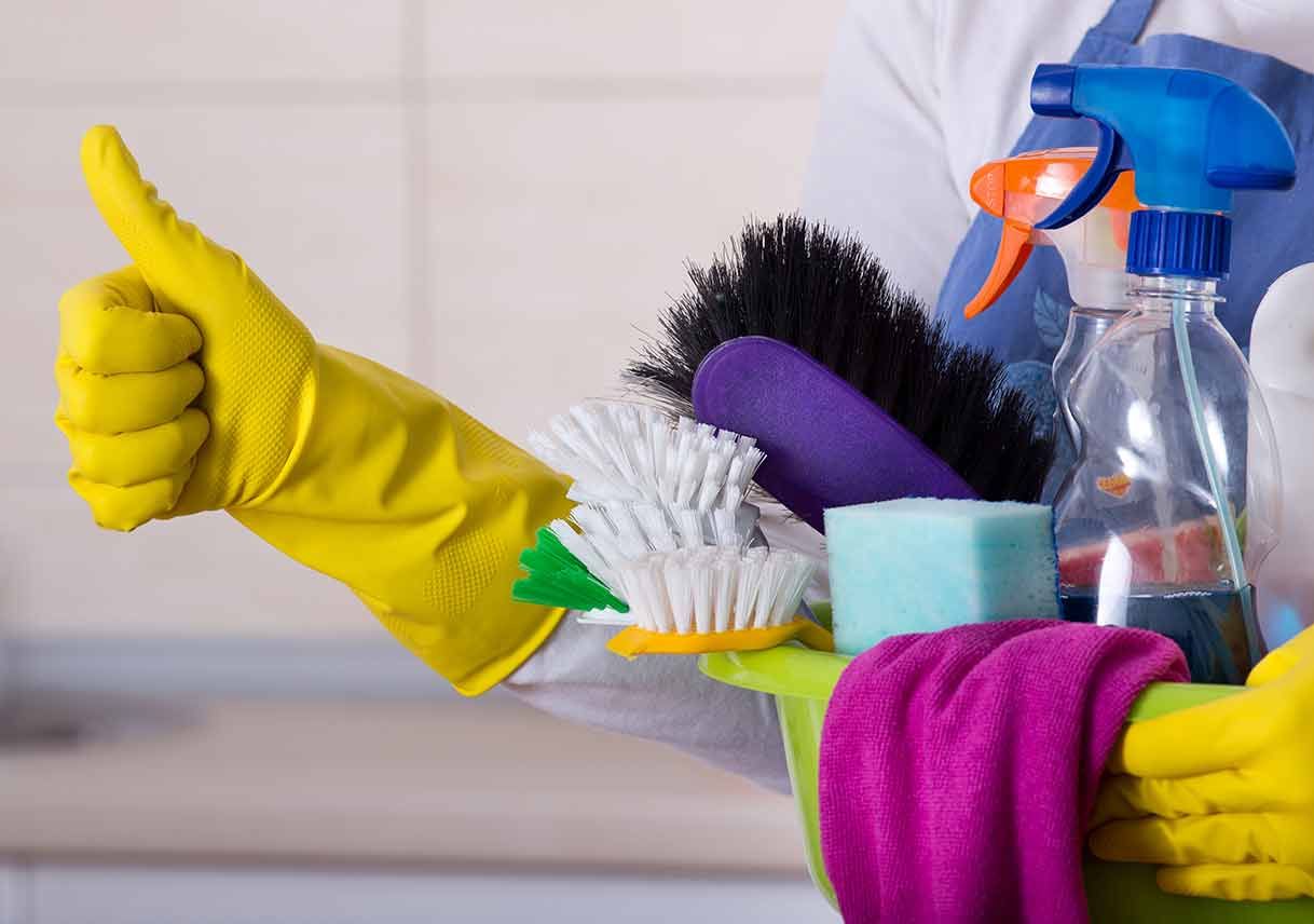 A person in yellow cleaning gloves gives a thumbs-up while holding a basin filled with sponges, brushes, and spray bottles.