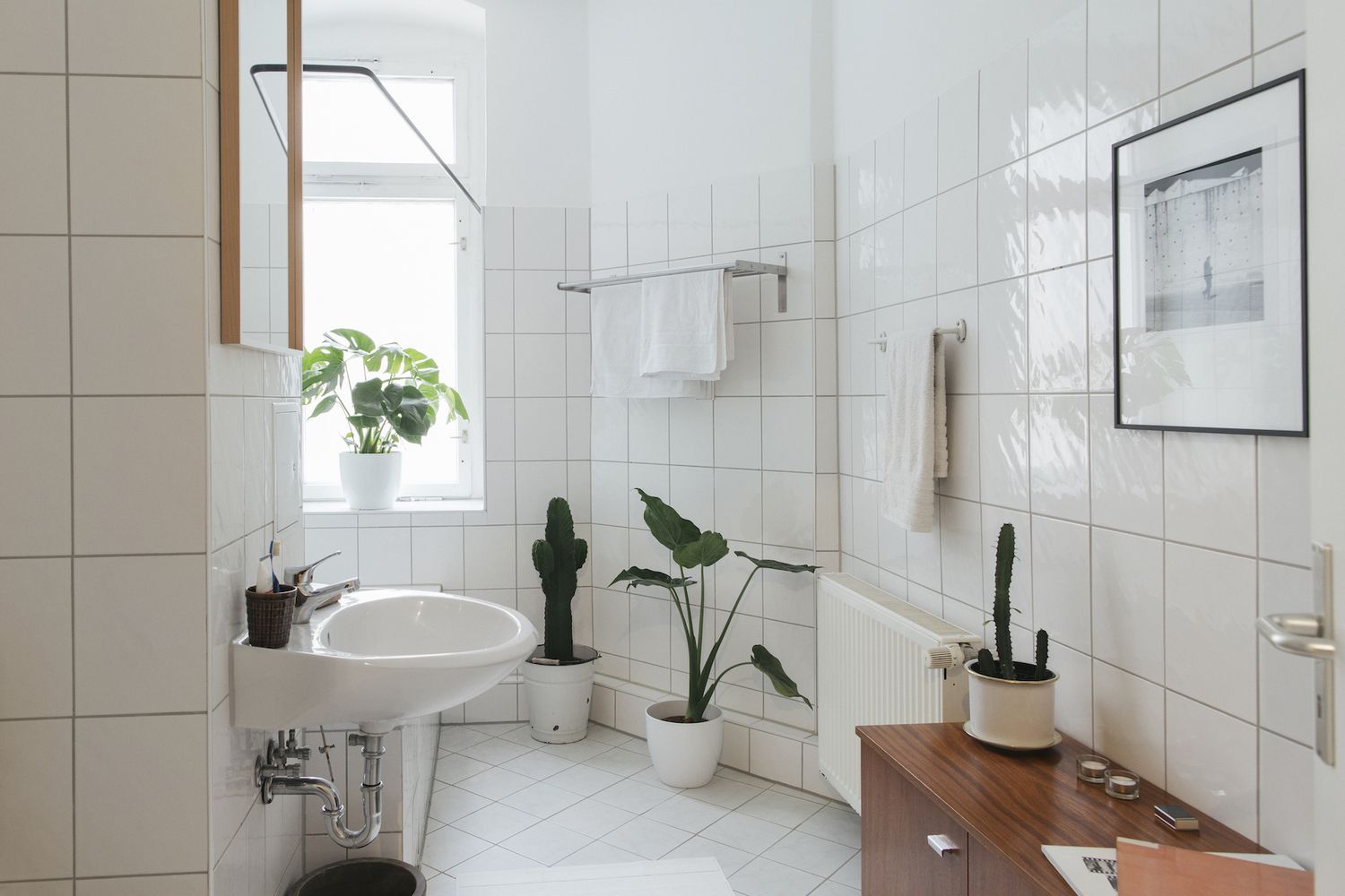 A bright, white-tiled bathroom featuring a pedestal sink, several potted green plants, and a wooden cabinet.