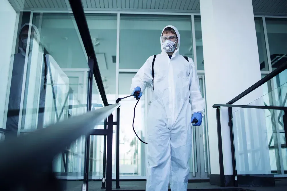 A person in a full white protective suit and face mask sprays disinfectant on a metal railing in a modern building.