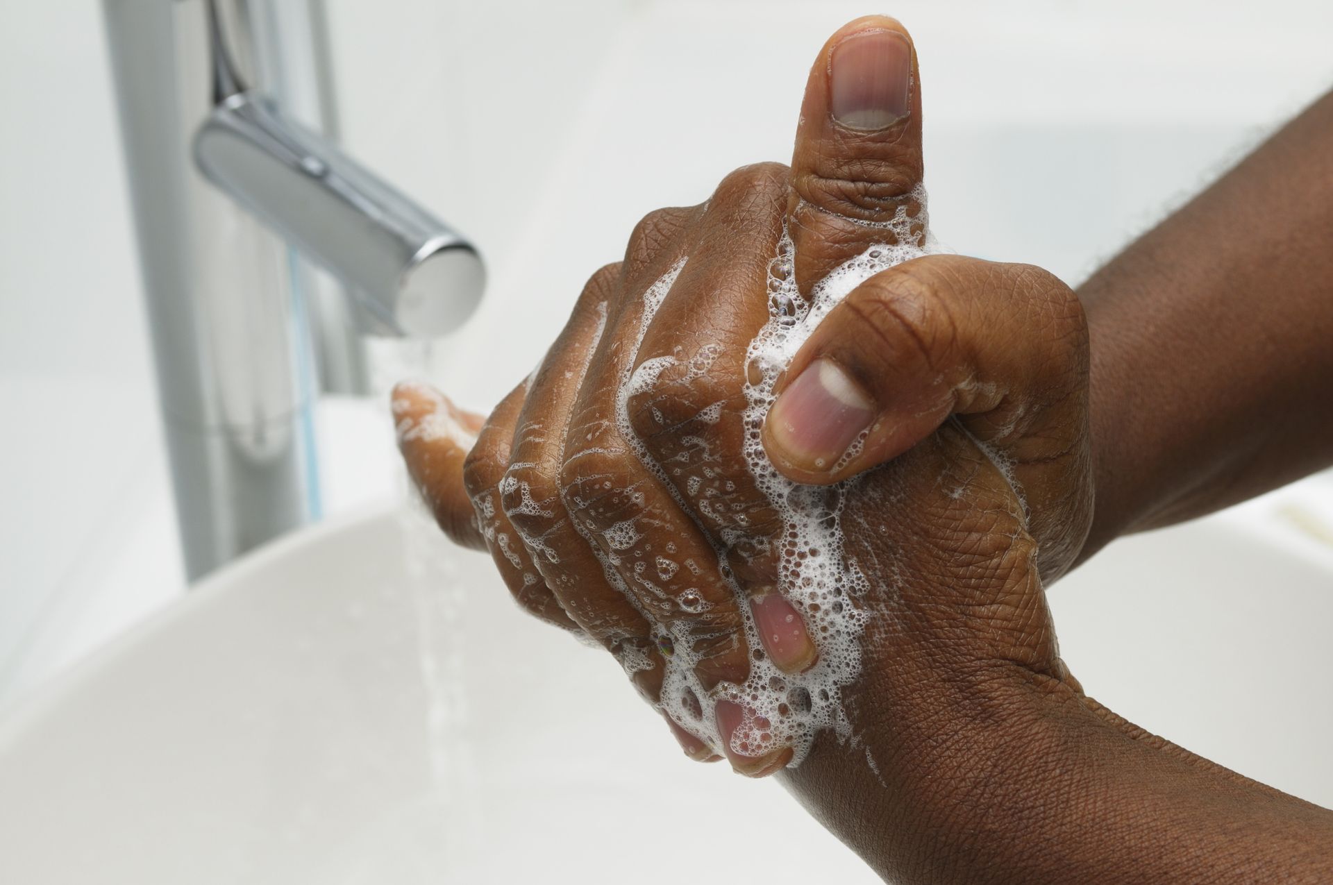 Hands lathered with white soap foam are being rubbed together under a running faucet at a sink.