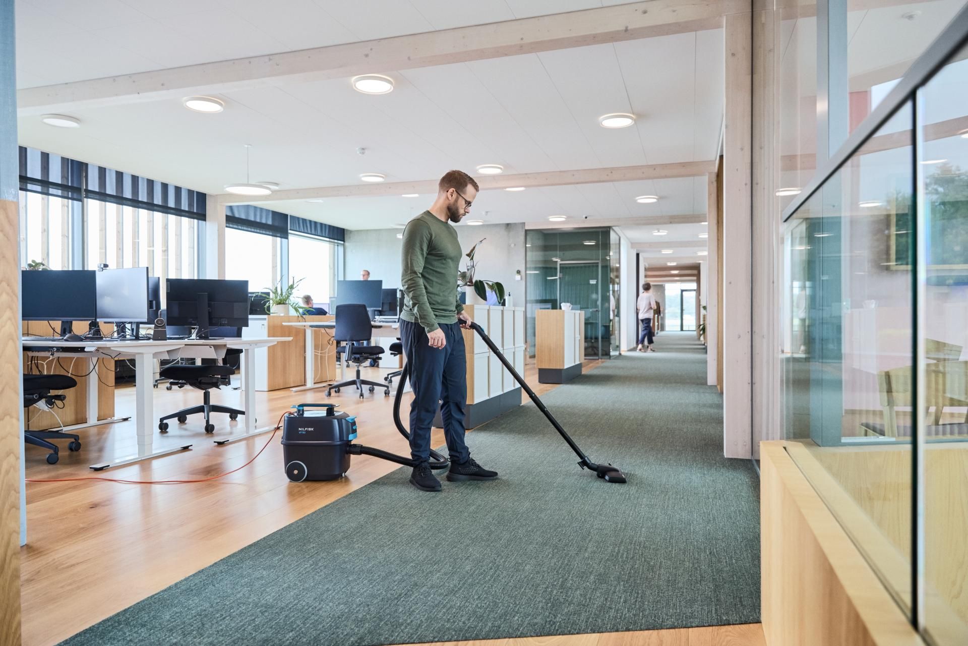 A person in an office workspace vacuums the green carpeted floor while standing between desk areas and a hallway.