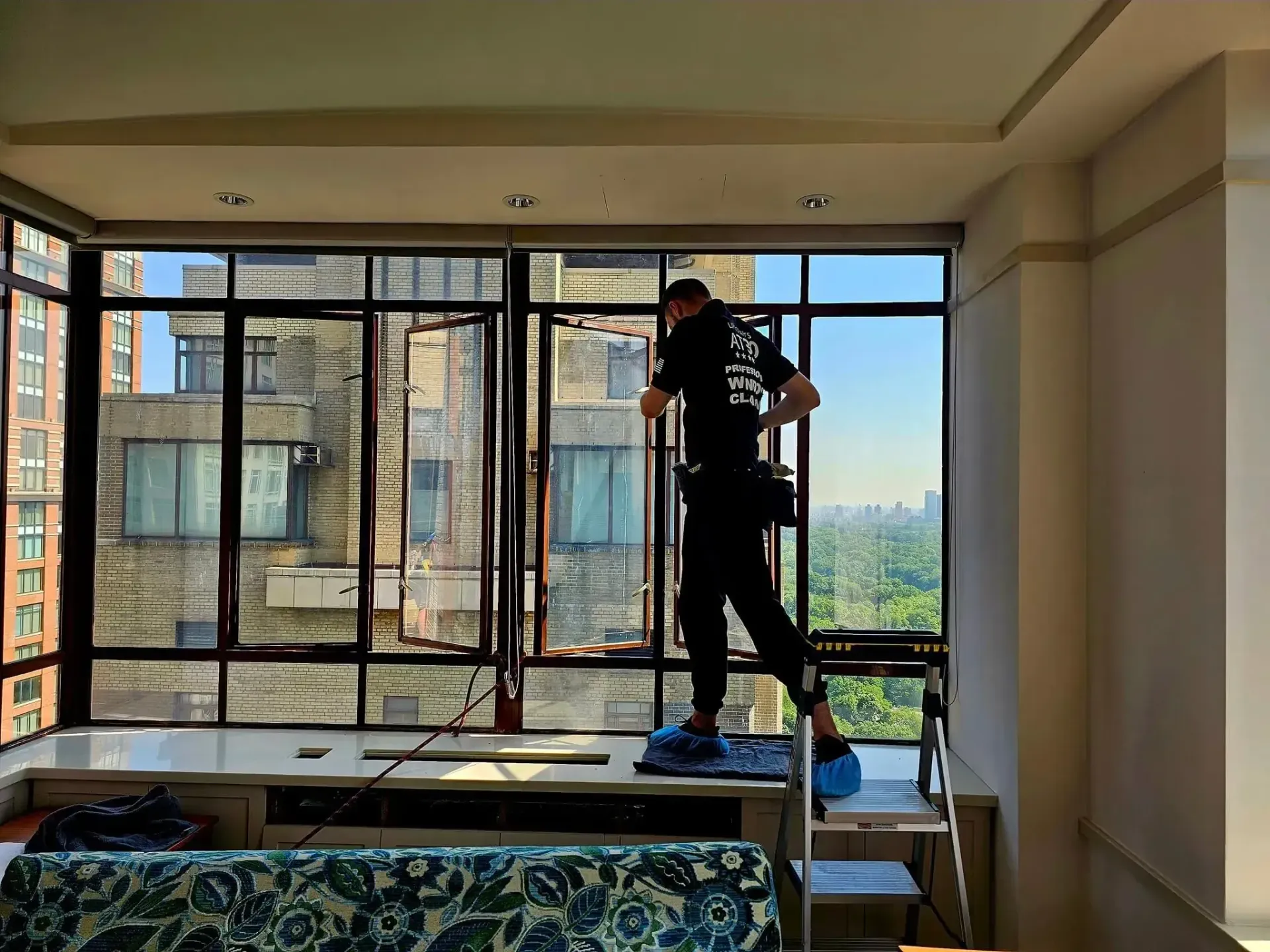 A worker in dark clothing stands on a small ladder, cleaning large apartment windows overlooking a park and city skyline.