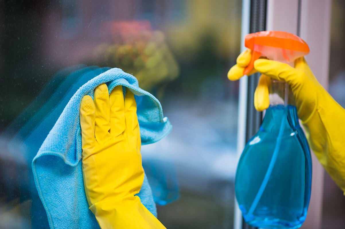 Hands wearing yellow rubber gloves cleaning a glass window with a blue cloth and a spray bottle of blue liquid.