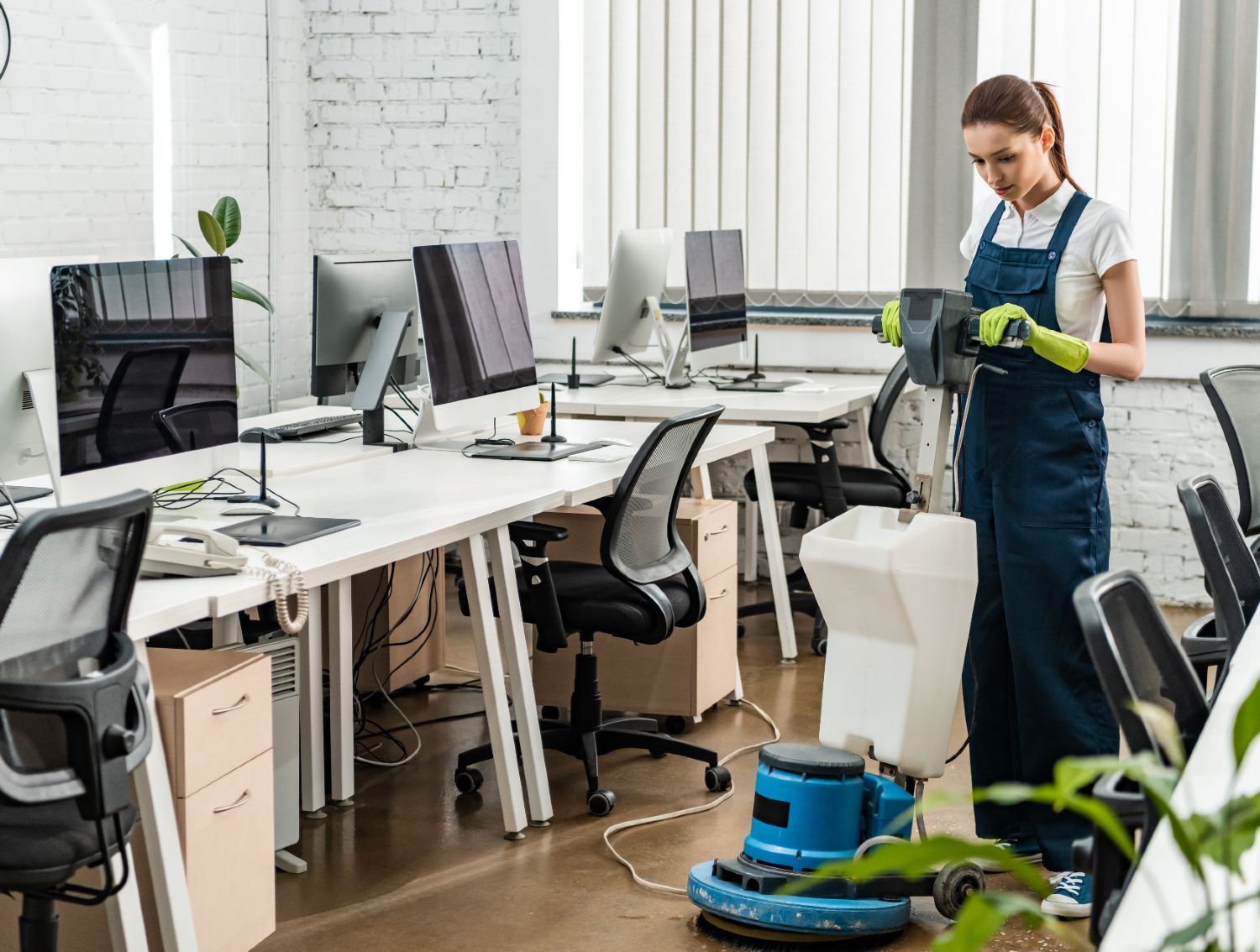 A cleaner in blue overalls and gloves operates a floor scrubber in a modern, sunlit open-plan office.