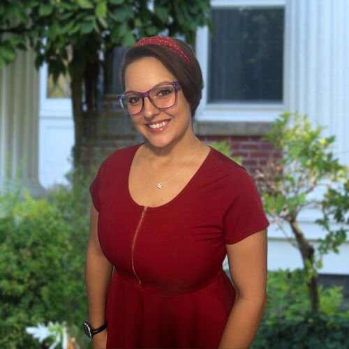 A woman wearing glasses and a red dress is standing in front of a house.