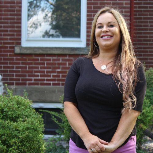 A woman in a black shirt and pink shorts is smiling in front of a brick building