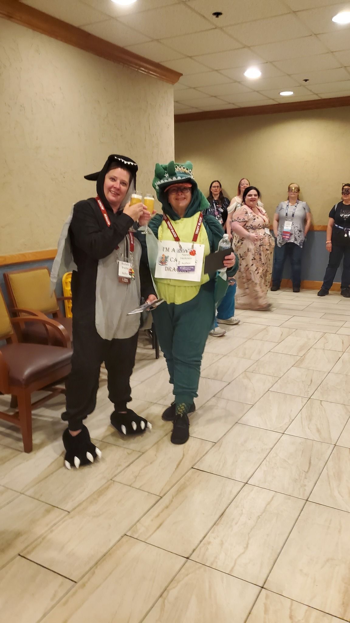 Two people in costumes, dragon and dinosaur, pose with drinks at an event.