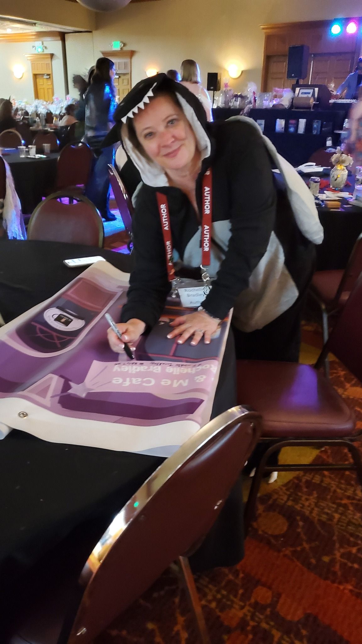Woman in shark hoodie signing merchandise at a table; convention setting, many people in background.