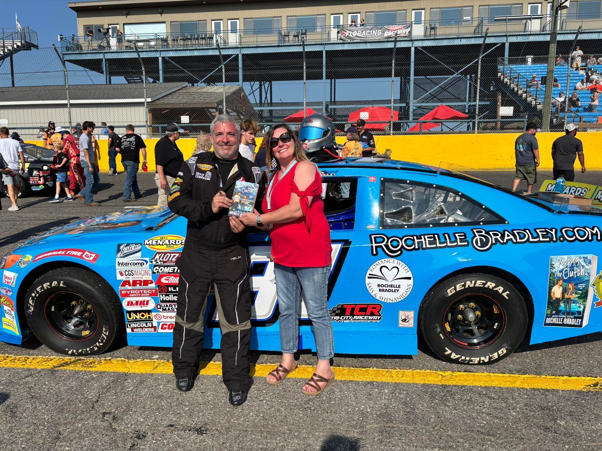 Man and woman with trophy in front of a blue race car at a track.