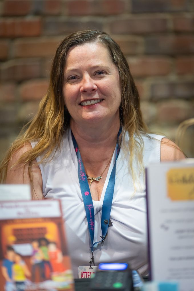 Woman with long hair, white top, smiling, with lanyard, sitting at a table.