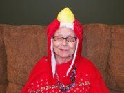 An elderly woman is sitting on a couch wearing a red parrot costume.