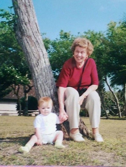A smiling older woman in a red shirt and khakis sits near a tree holding the hand of a baby wearing a white onesie.
