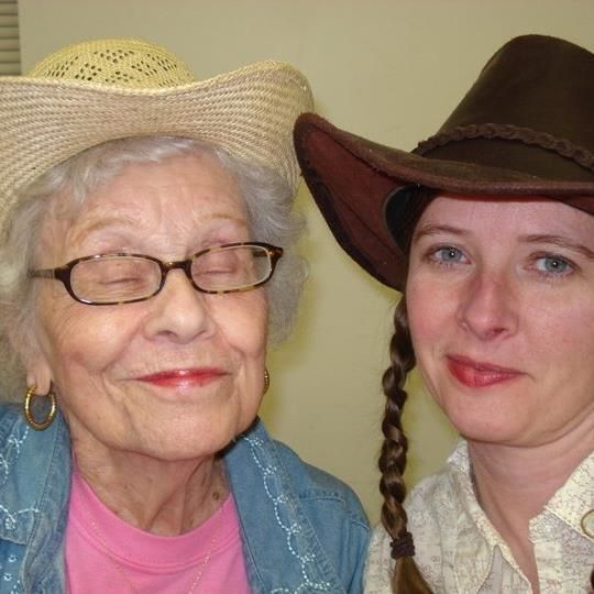 An elderly woman and younger woman in cowboy hats pose together. The elderly woman smiles, wearing glasses and a straw hat. The younger woman has a braided pigtail and wears a brown hat.