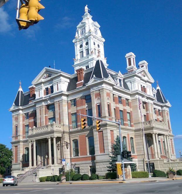 A large, ornate brick courthouse with a clock tower against a bright blue sky. Traffic lights are visible in front.