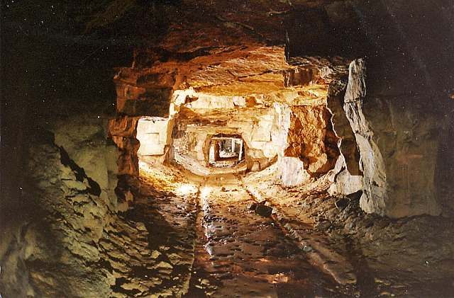 A dark, layered tunnel with rough, brown rock walls leading to a distant light source.