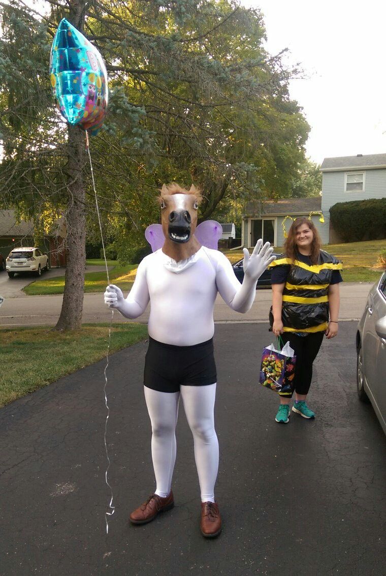 A person in a horse head costume, holding a balloon, waves next to someone dressed as a bee on a driveway.