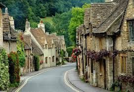 Stone buildings line a winding road in a quaint village with green foliage.