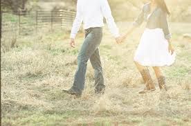 A couple walks hand-in-hand through a field. The woman wears a white dress and boots, the man jeans and a white shirt.