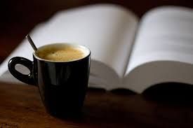 A black coffee mug with a spoon sits in front of an open book on a wooden surface.