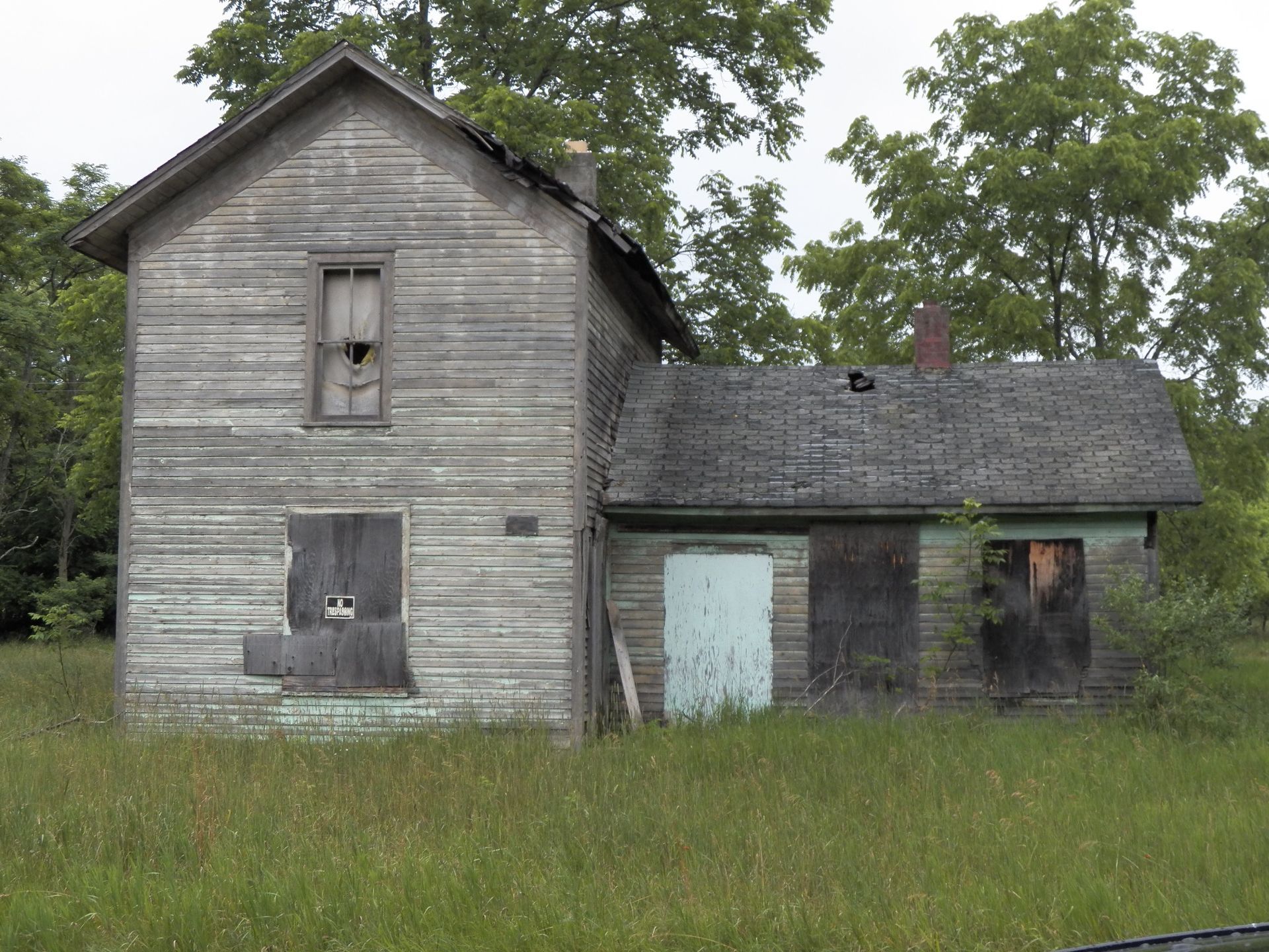 Dilapidated, two-story wooden house with boarded-up windows and doors in a field of tall grass.