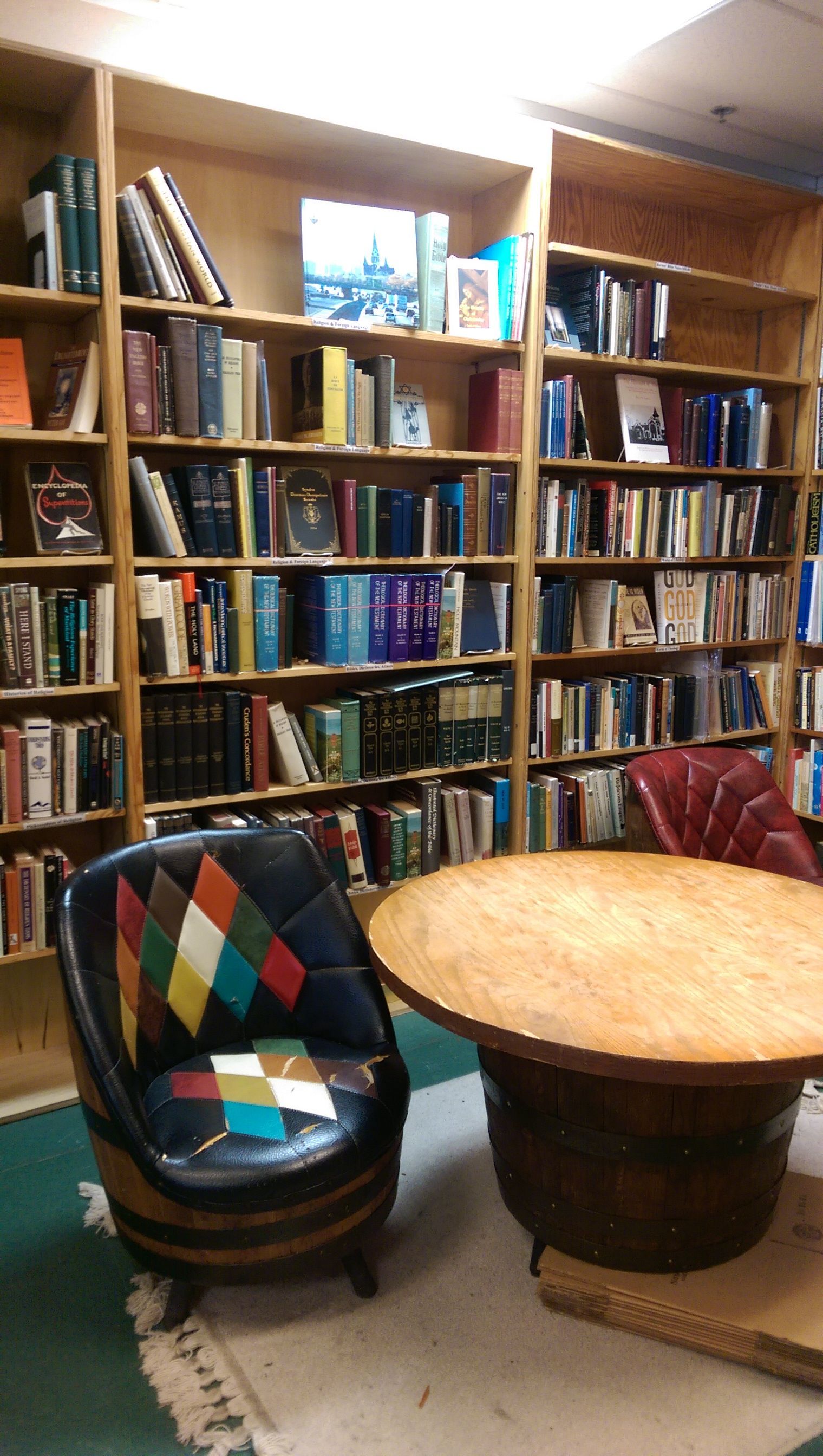 A cozy bookstore interior with tall wooden bookshelves filled with books, a round wooden table, and a barrel chair with a colorful patchwork back.