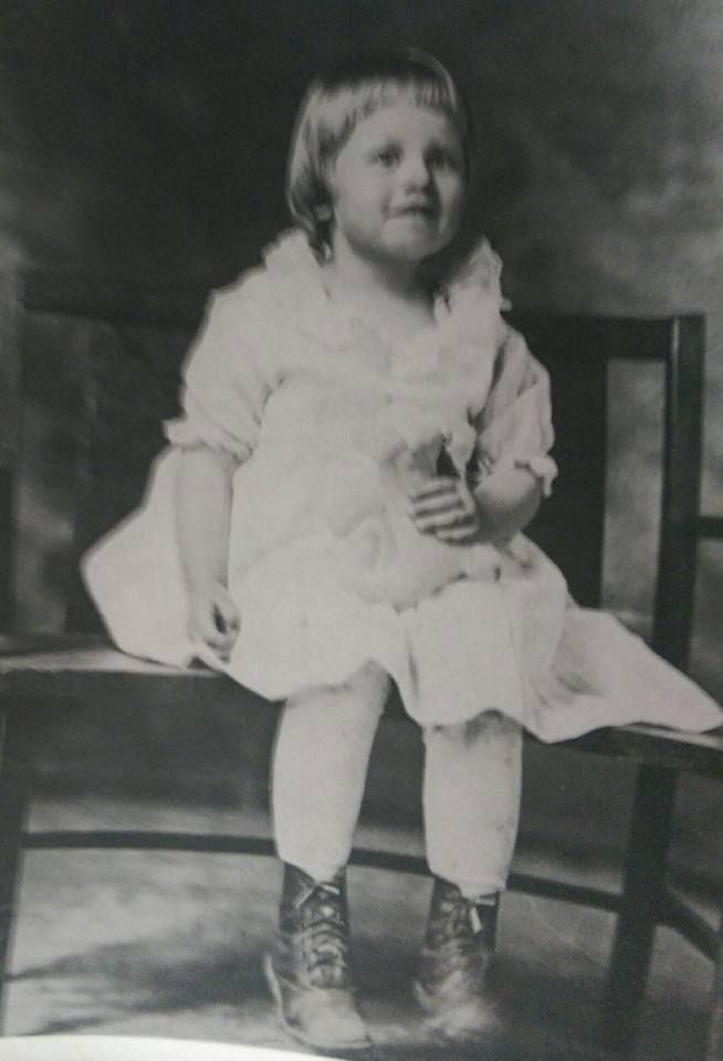 A young child in a frilly white dress and boots sits on a bench, holding a small item, with a soft smile.