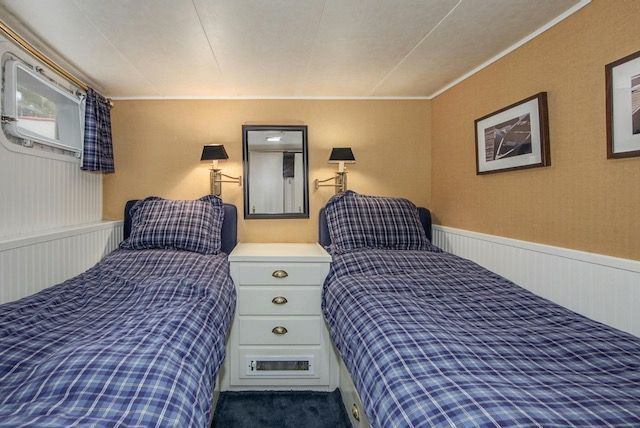 Twin beds in a small room with blue plaid bedding, flanked by nightstands, lamps, and framed art.