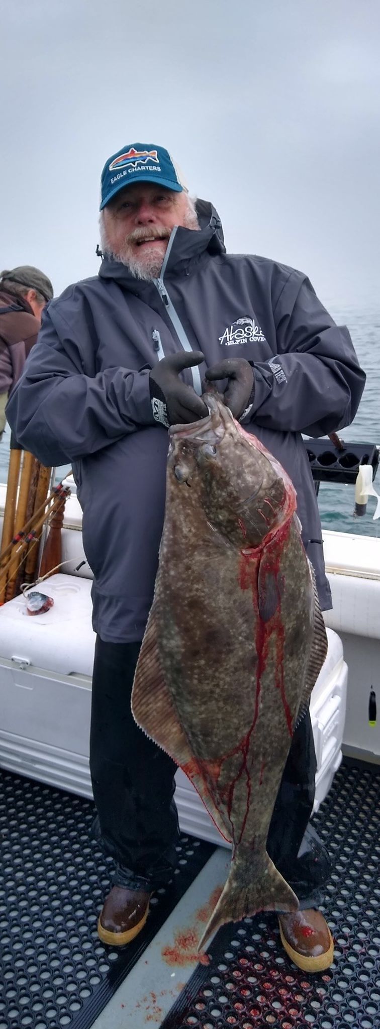 Man holding a large halibut on a boat; gray day.