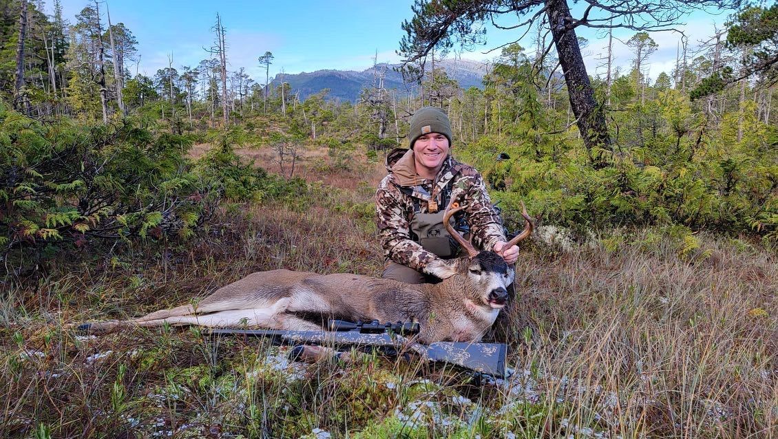 Hunter in camouflage kneels beside a deer on the ground, rifle nearby, in a wooded area.