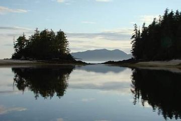 Still water reflects forest islands and distant mountains under a cloudy sky.