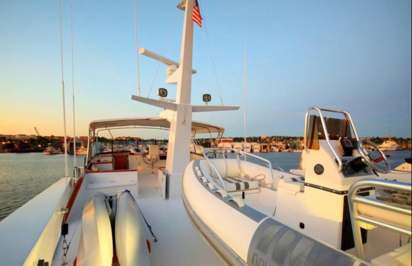 A luxury yacht's deck with a dinghy, equipment, and a city harbor in the background at sunset.