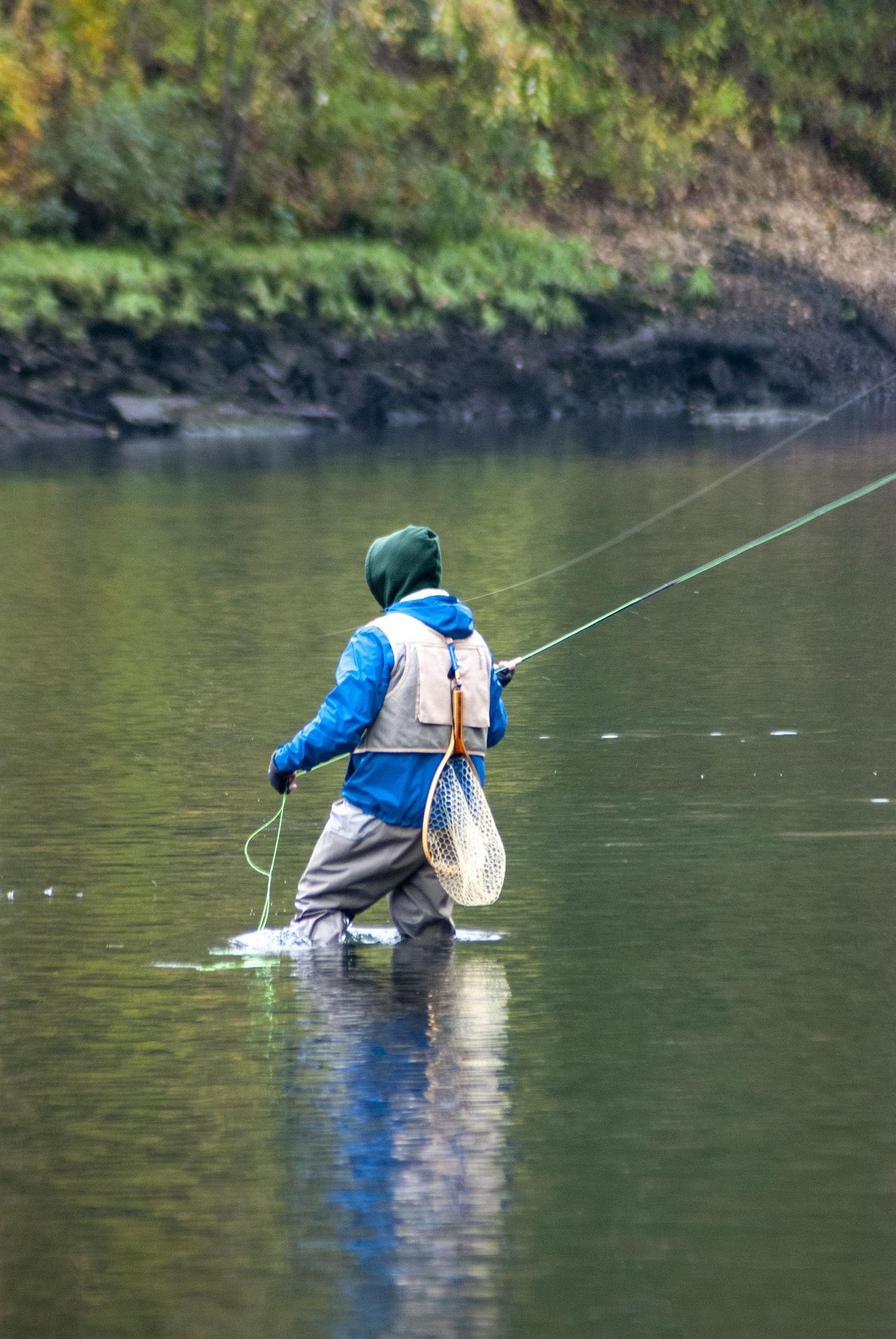 Fisherman wading in river, casting a line. Wearing blue jacket, tan waders, and green hood. Net attached.