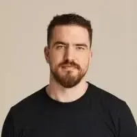 A man with a beard is wearing a black t-shirt and looking at the camera.