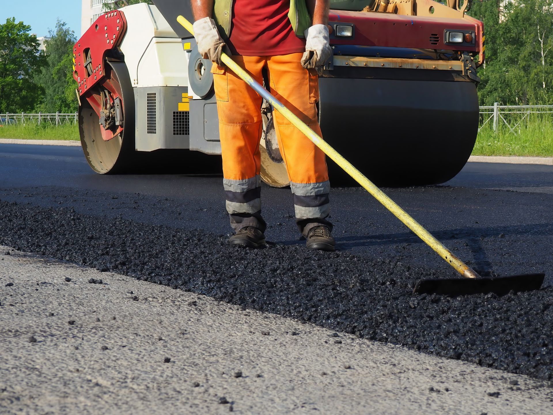A worker rakes asphalt while a roller compacts pavement near trees and a fence.