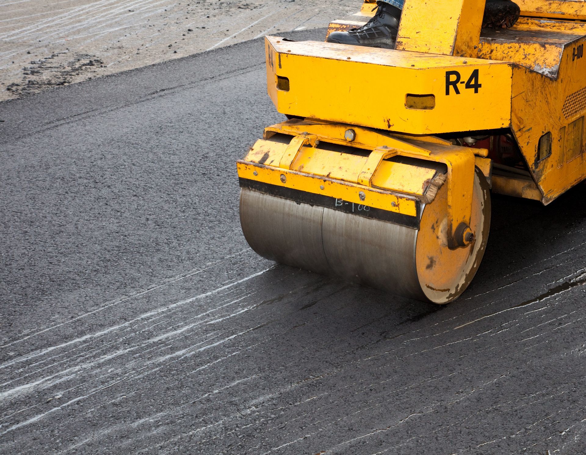 A yellow asphalt flattener flattens a road of freshly placed asphalt on the ground.