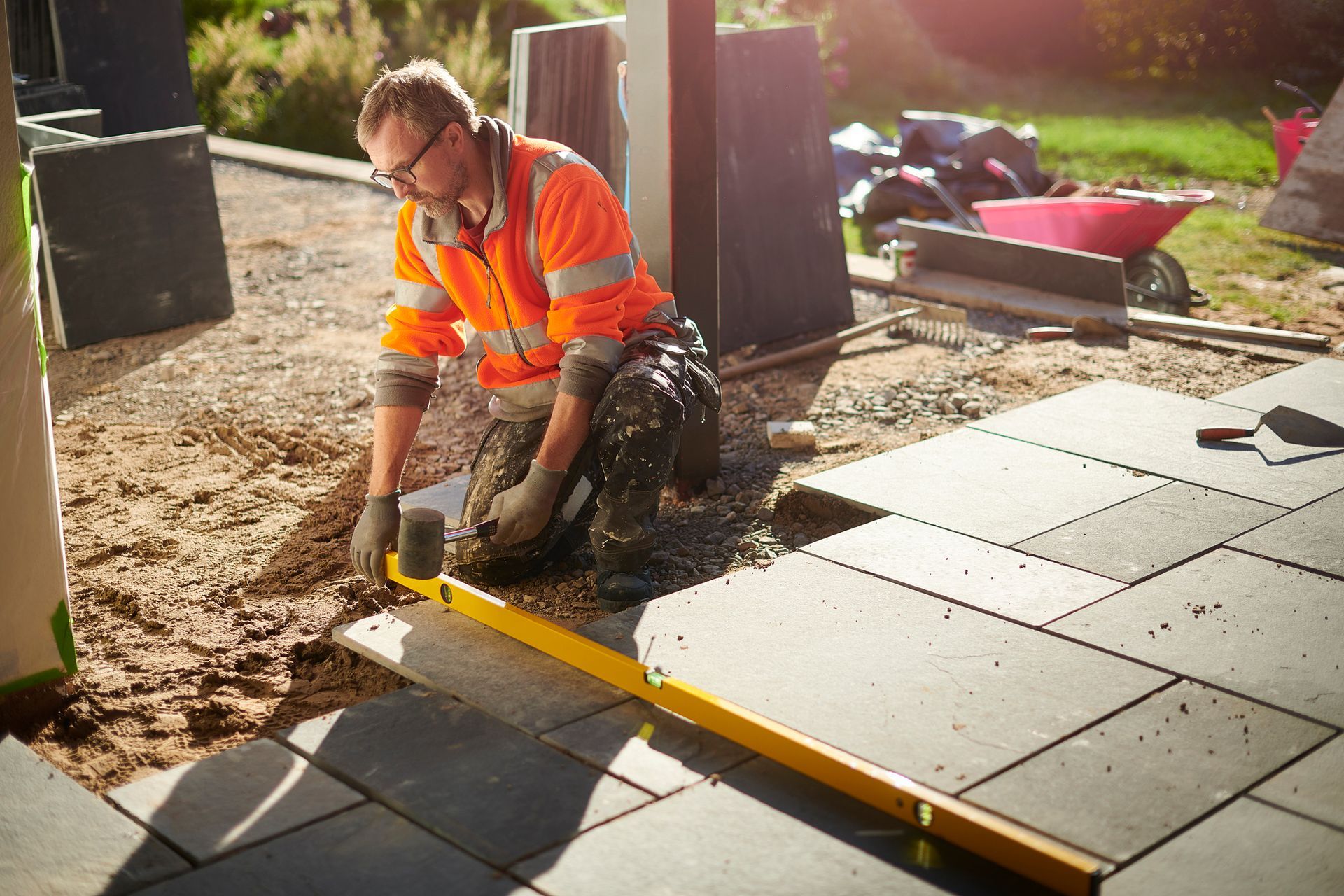 Construction worker kneels, leveling paving stones outdoors, wearing orange safety vest.