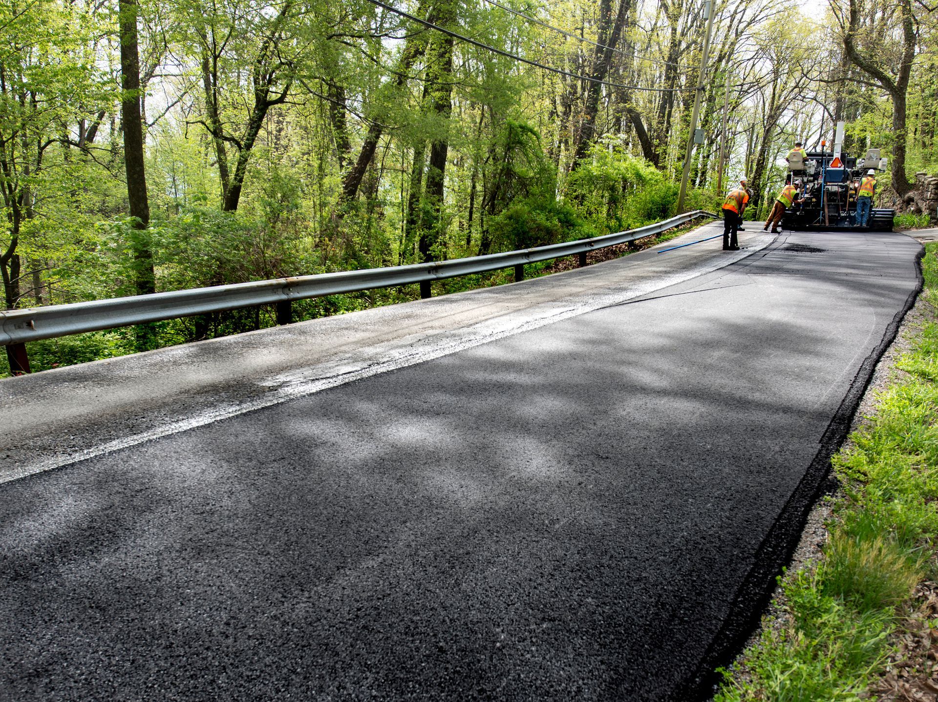 Asphalt road being paved, with machinery and workers. Road is beside a guardrail and trees.