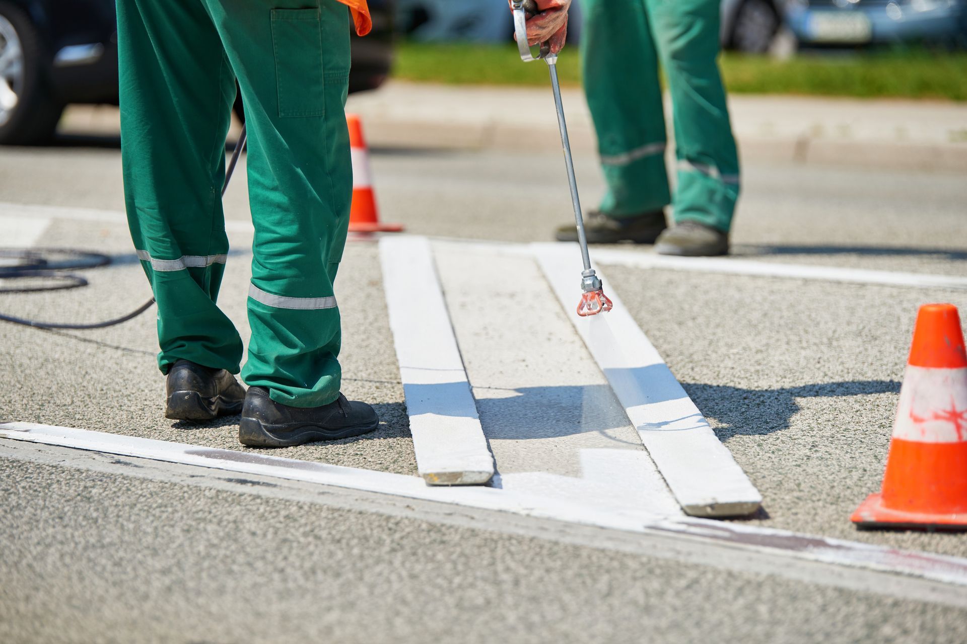 Two workers in green painting a white crosswalk on asphalt, with cones and a car visible.