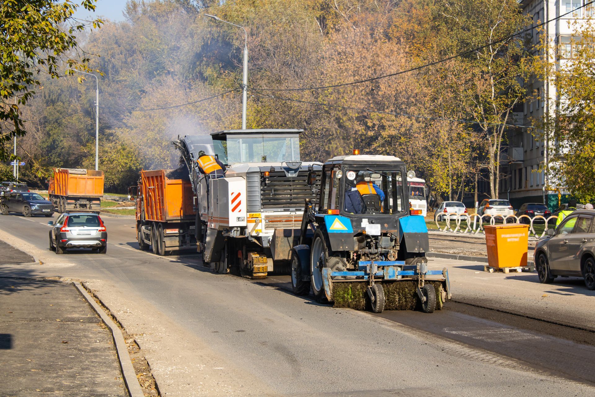 Road paving machinery resurfacing a street with vehicles and buildings in the background.