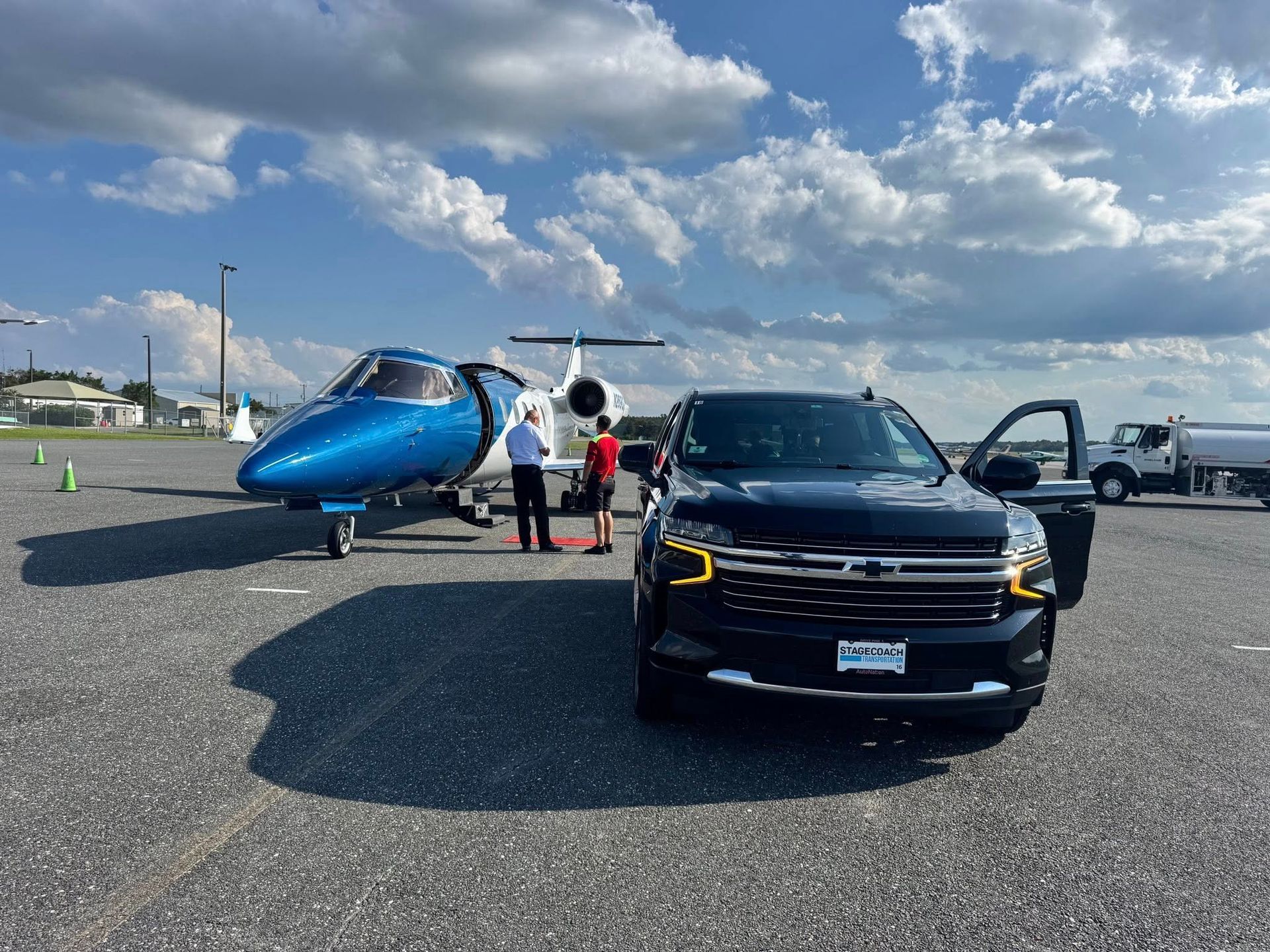 A black SUV is parked on an airfield next to a blue and white private jet, with two people standing near the aircraft.
