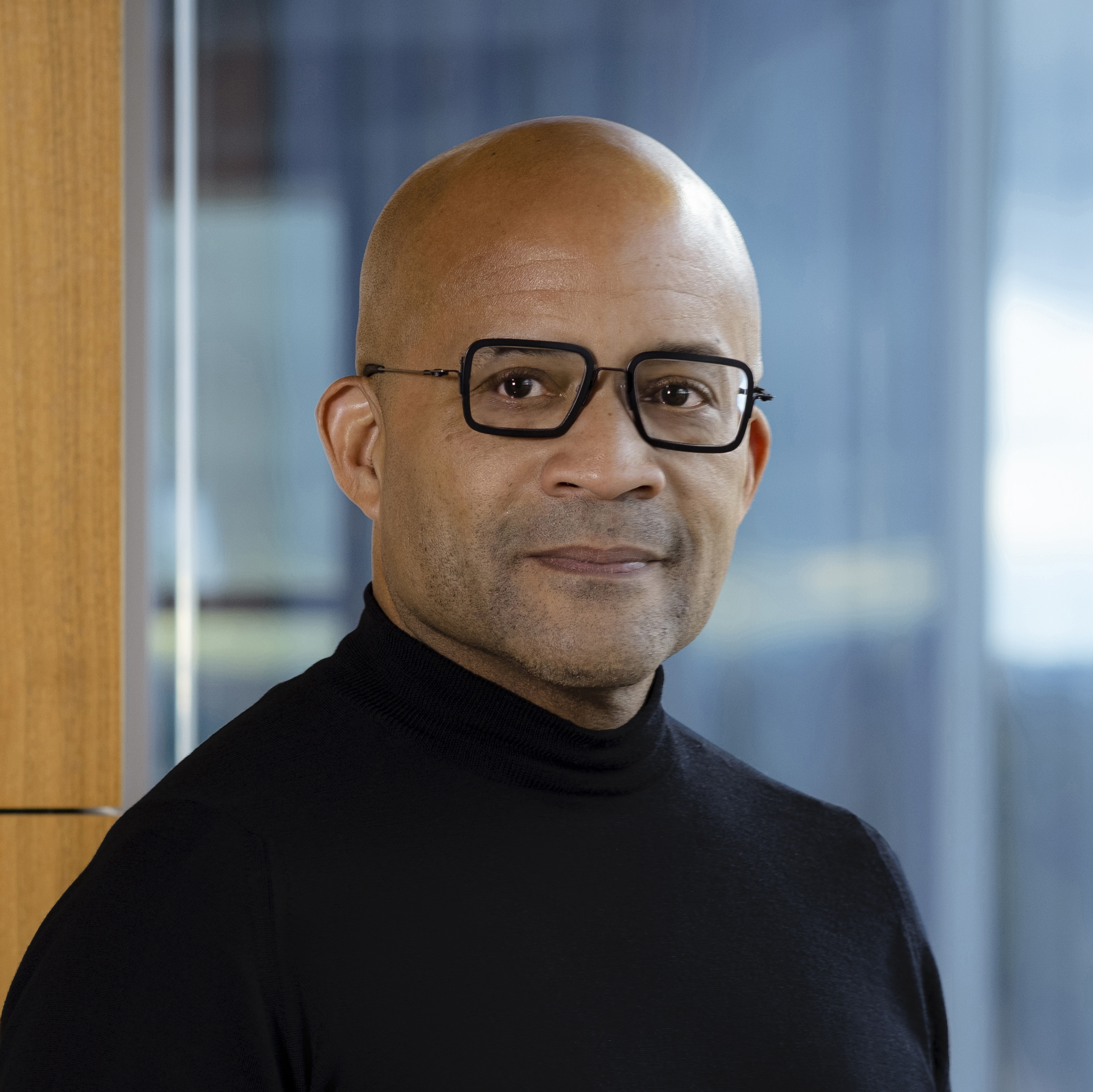 A bald man wearing glasses and a black turtleneck is standing in front of a window.